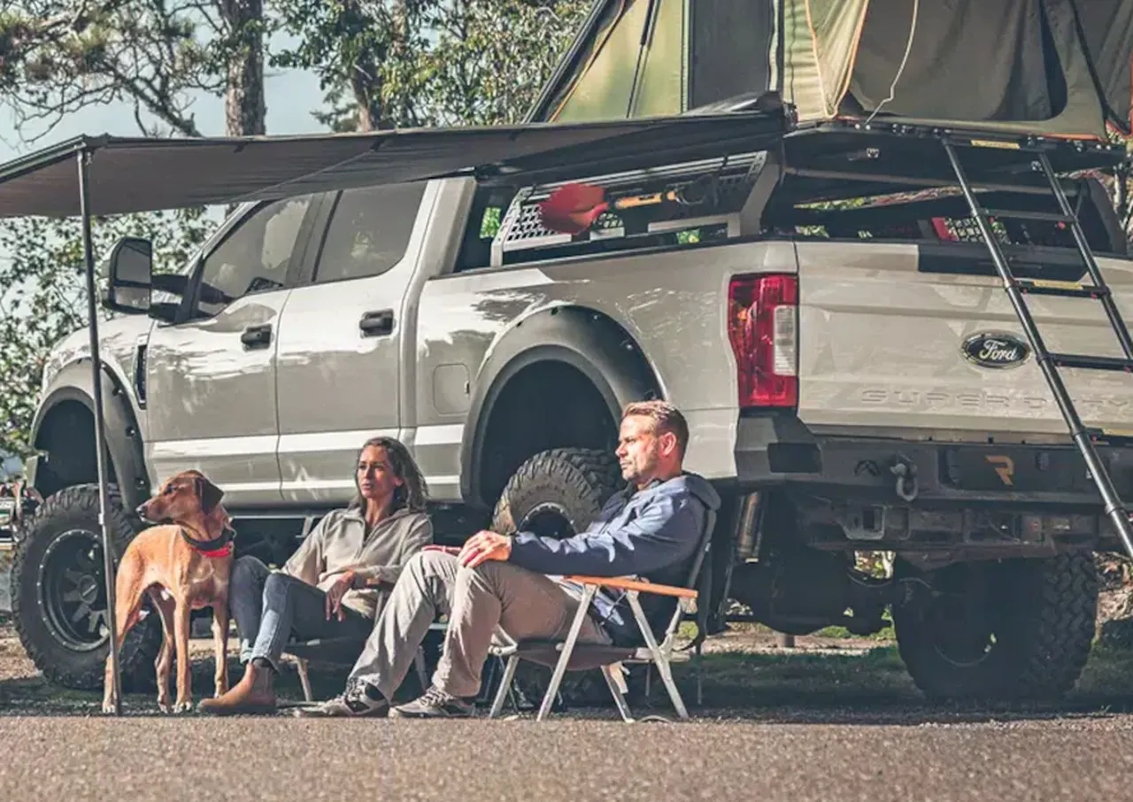 People relaxing under a truck-turned-campsite with a dog nearby during outdoor camping.