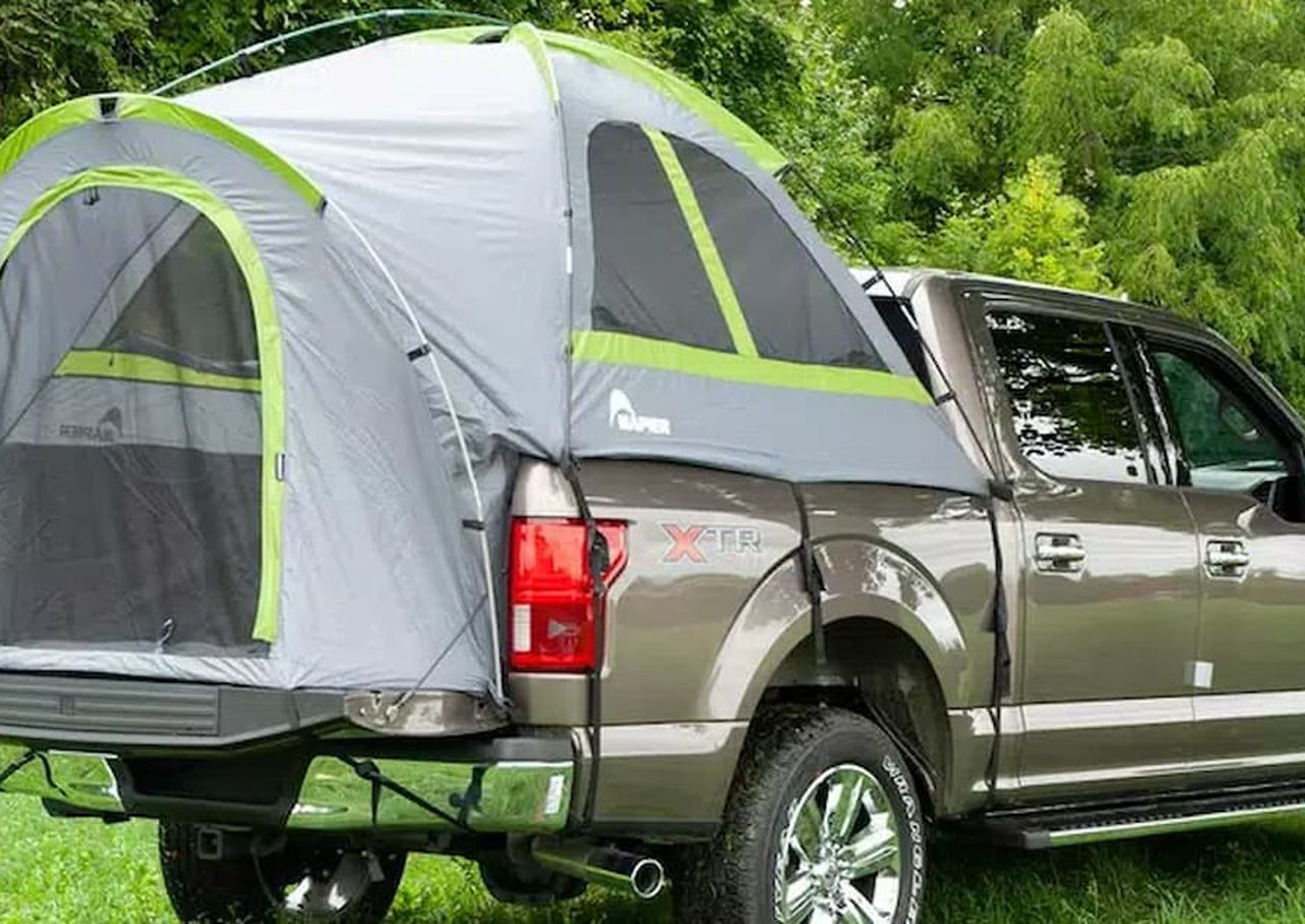 Pickup truck with a pop-up tent attached in a grassy, wooded area.