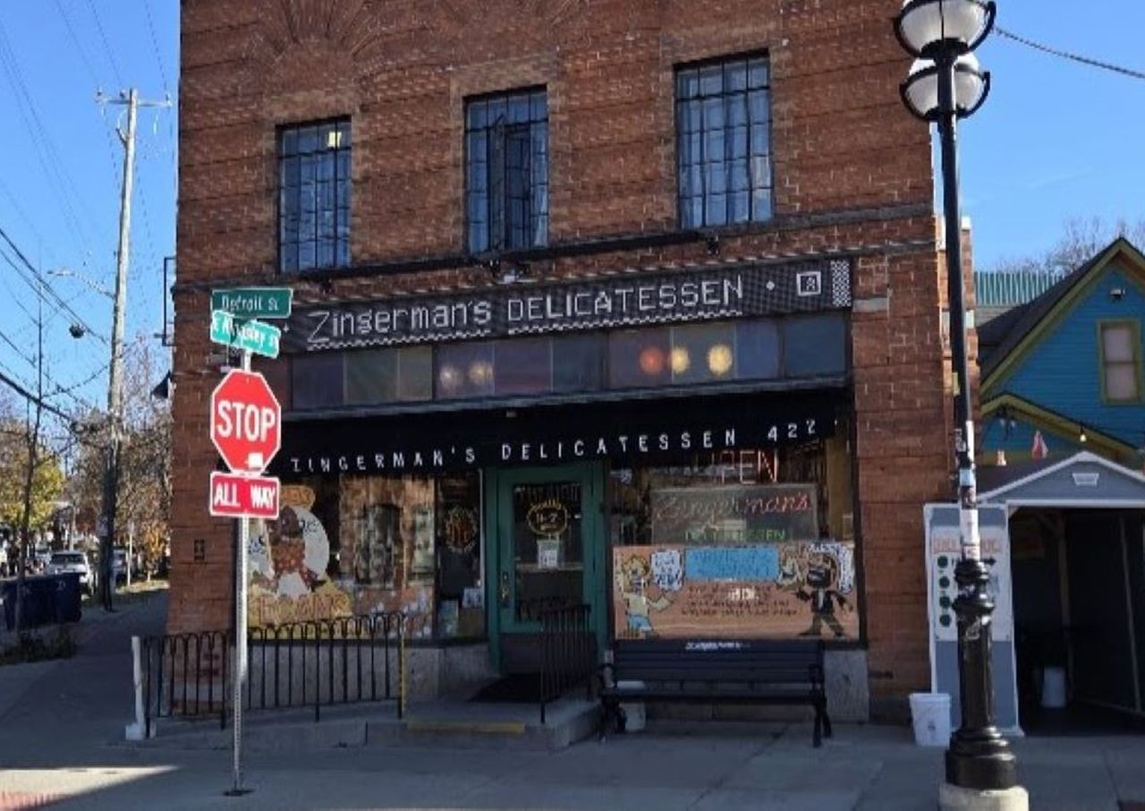 Brick building with "Zingerman's Delicatessen" sign, street signs, and a stop sign in front. Clear blue sky and small surrounding buildings.