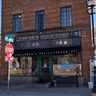 Brick building with "Zingerman's Delicatessen" sign, street signs, and a stop sign in front. Clear blue sky and small surrounding buildings.