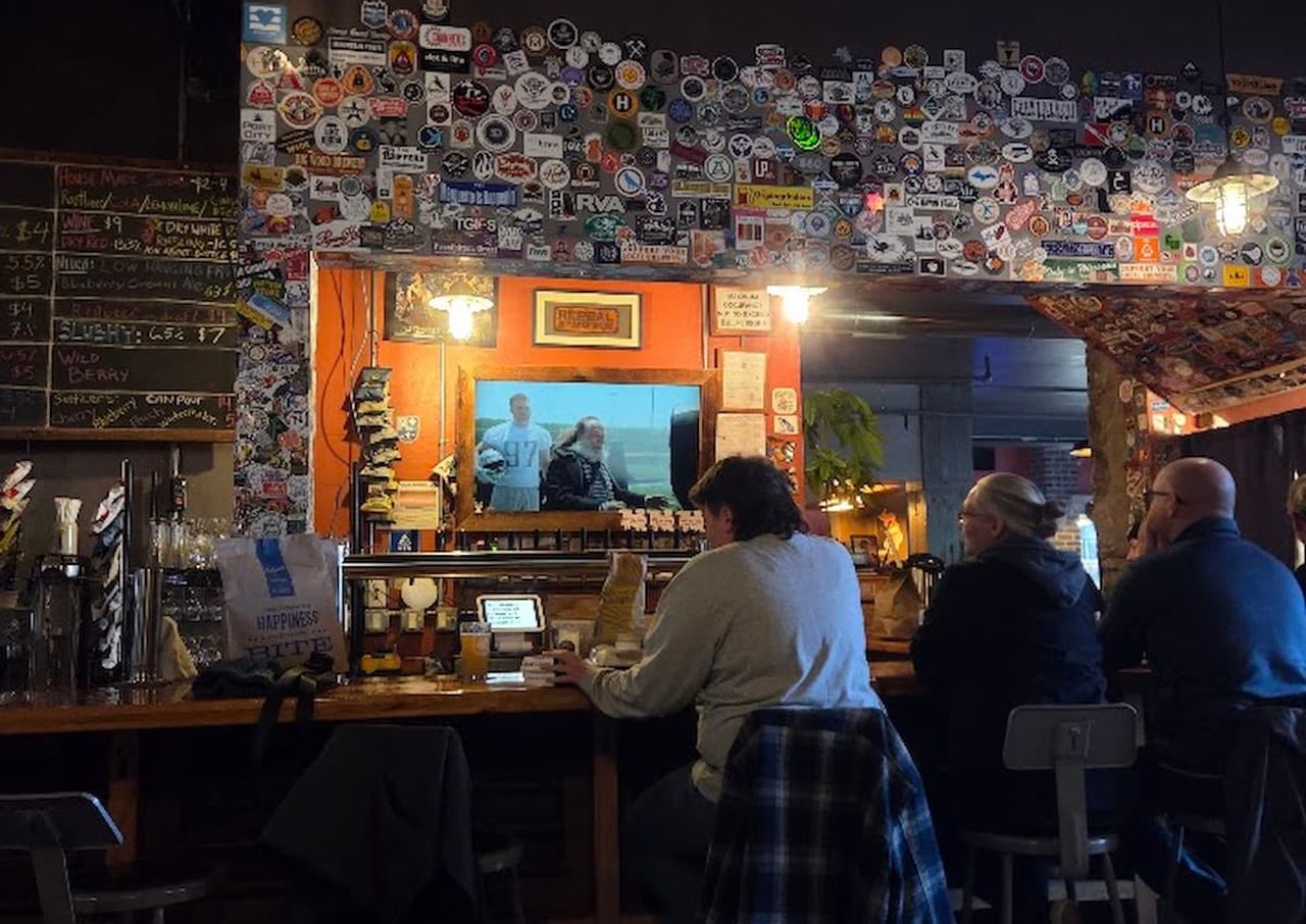 People sitting at a bar at Ore Dock Brewing Company with a wall covered in stickers and a small TV screen displaying a show.