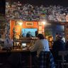 People sitting at a bar at Ore Dock Brewing Company with a wall covered in stickers and a small TV screen displaying a show.