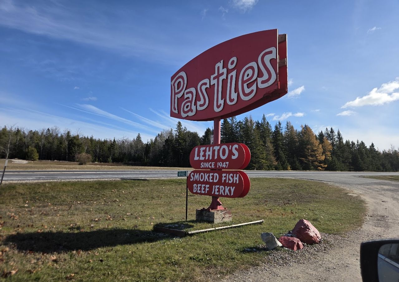 A roadside sign for "Pasties" with additional signs for "Lehto's," "Smoked Fish," and "Beef Jerky" on a grassy area under a blue sky.