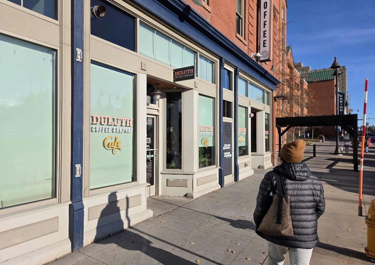 Person in a winter coat walking past Duluth Coffee Company, a cafe with large windows on a sunny street.