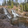 Waterfalls cascade over rocky ledges into a calm pool, surrounded by trees and a few people observing from the top.