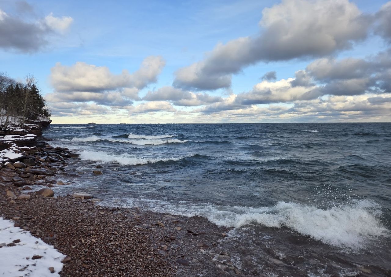 Rocky shoreline of Lake Superior with gentle waves under a partly cloudy sky, bordered by snow-dusted trees on the left.