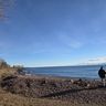 Person walking along a rocky shoreline with a clear blue sky and calm water, surrounded by bare trees and sparse vegetation.
