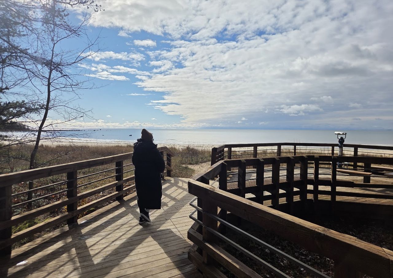 Person in a coat walking on a wooden boardwalk towards a lake under a partly cloudy sky.
