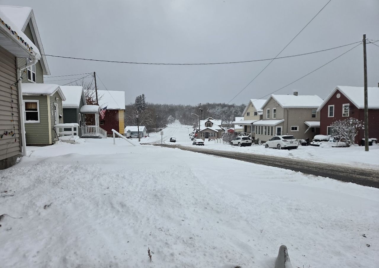 Snow-covered suburban street with houses on both sides, overcast sky, and a few cars parked along the road.
