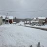 Snow-covered suburban street with houses on both sides, overcast sky, and a few cars parked along the road.