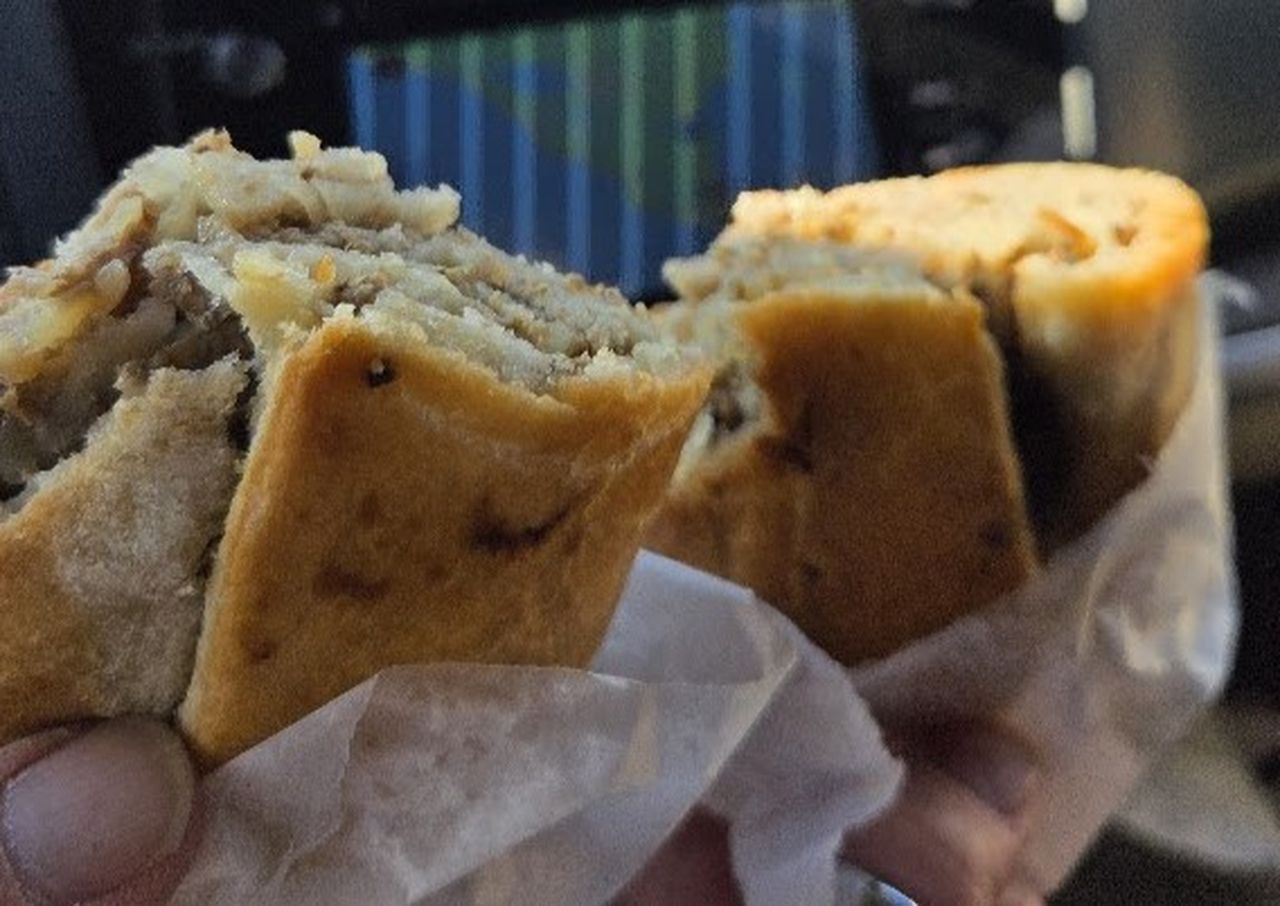 Two hands holding halves of a savory pastry filled with meat and cheese, wrapped in paper, with a blurred background of a car interior.